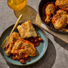A plate of braised chicken with beans and bread is presented alongside a cast iron skillet containing more of the same dish, suggesting a hearty and flavorful meal.