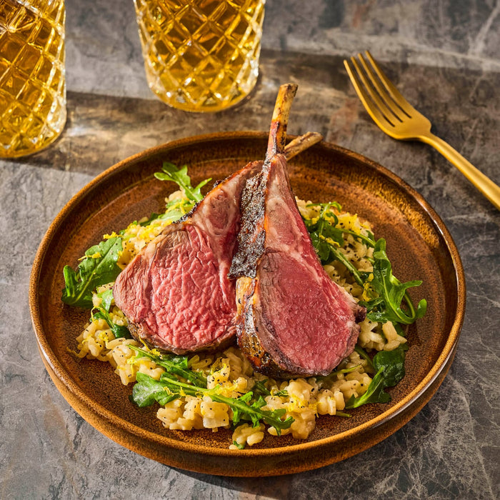 A plate of perfectly cooked lamb chops served over a bed of greens and grains, accompanied by a golden fork and glasses of beer.