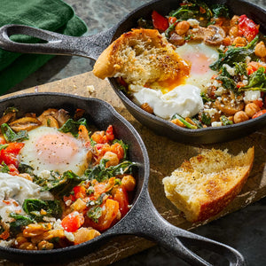 Two cast iron skillets hold a vibrant breakfast dish of chickpeas, spinach, tomatoes, feta cheese, and a sunny-side-up egg, garnished with toasted bread.