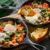 Two cast iron skillets hold a vibrant breakfast dish of chickpeas, spinach, tomatoes, feta cheese, and a sunny-side-up egg, garnished with toasted bread.