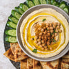 A bowl of hummus with chickpeas, olive oil, and spices, surrounded by pita bread and cucumber slices.