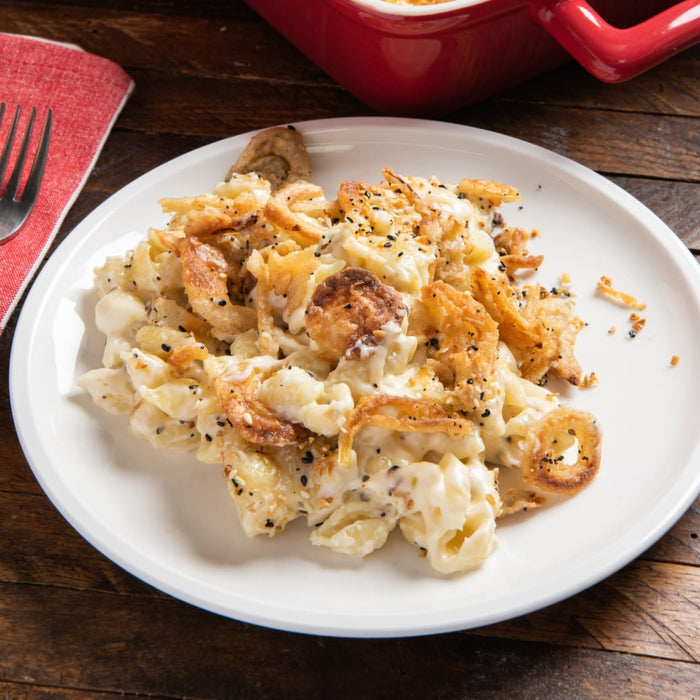 A creamy serving of macaroni and cheese is topped with crispy fried onions and a generous sprinkle of Mccormick Everything Bagel Seasoning Assorted Form 4.8 Oz on a white plate.
