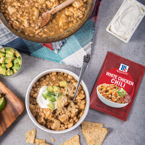 A bowl of white chicken chili garnished with avocado and sour cream sits next to a packet of McCormick White Chicken Chili seasoning mix.
