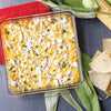 A square glass baking dish filled with corn dip, topped with crumbled cheese and herbs, served with tortilla chips.