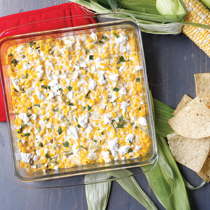 A square glass baking dish filled with corn dip, topped with crumbled cheese and herbs, served with tortilla chips.