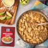 A packet of McCormick Spicy Chicken Chili mix is displayed next to a pan of cooked chili, shredded cheese, diced avocado, and lime wedges.