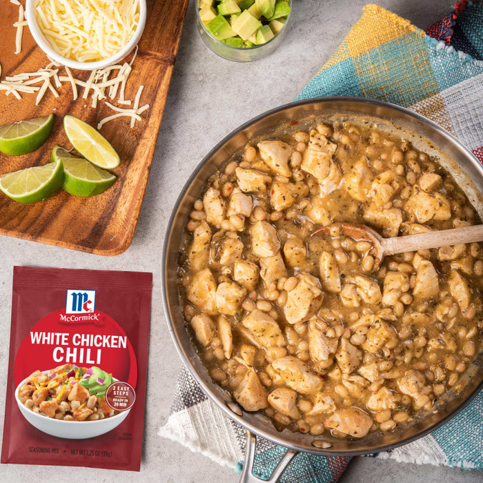 A packet of McCormick Spicy Chicken Chili mix is displayed next to a pan of cooked chili, shredded cheese, diced avocado, and lime wedges.