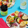 A jar of I Beg Your Garden veggie topping seasoning next to a plate of grilled vegetables on skewers.