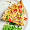 A slice of quiche with red peppers and asparagus on a white plate with a side of arugula.