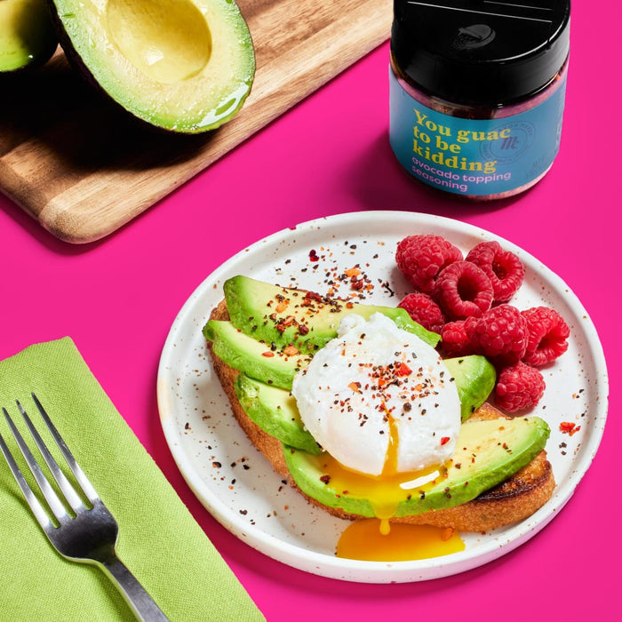A plate of avocado toast topped with a poached egg and raspberries, with a jar of You Guac to be Kidding avocado topping seasoning.