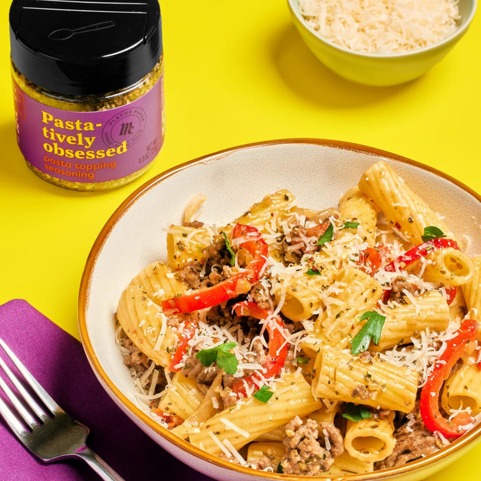 A bowl of pasta with ground beef, red peppers, and parmesan cheese, with a jar of Pasta-tively Obsessed seasoning in the background.