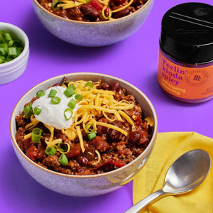 A bowl of chili topped with sour cream, shredded cheese, and green onions, next to a container of Flavor Maker Feelin' Kinda Spicy chili topping seasoning.