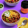 A bowl of chili topped with sour cream, shredded cheese, and green onions, next to a container of Flavor Maker Feelin' Kinda Spicy chili topping seasoning.