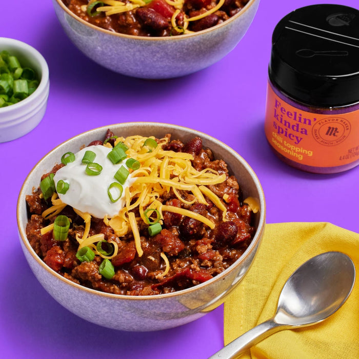 A bowl of chili topped with sour cream, shredded cheese, and green onions, next to a container of Flavor Maker Feelin' Kinda Spicy chili topping seasoning.