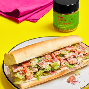 A sandwich with lettuce, tomato, salami, and cheese on a white plate with a jar of I'm Great in Bread sandwich topping seasoning.