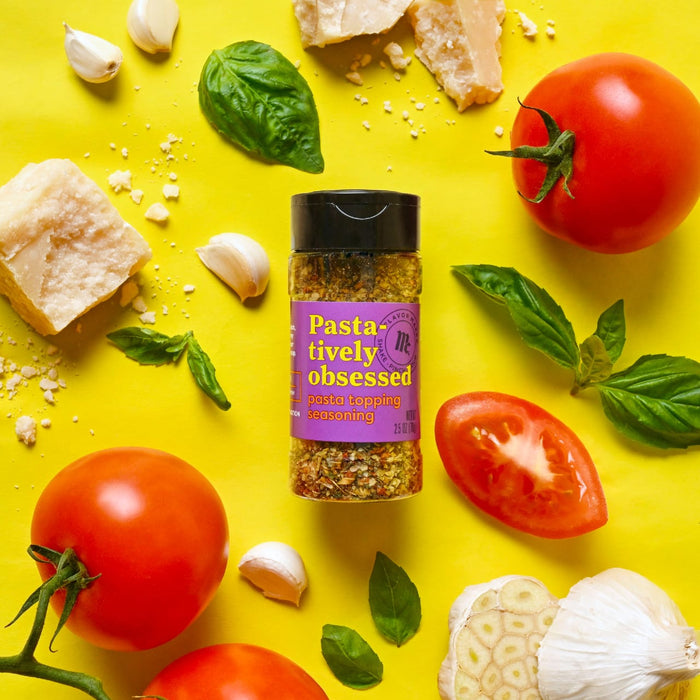 A jar of Flavor Makers Pasta-tively Obsessed pasta topping seasoning is surrounded by fresh ingredients like tomatoes basil garlic and parmesan cheese on a yellow background.