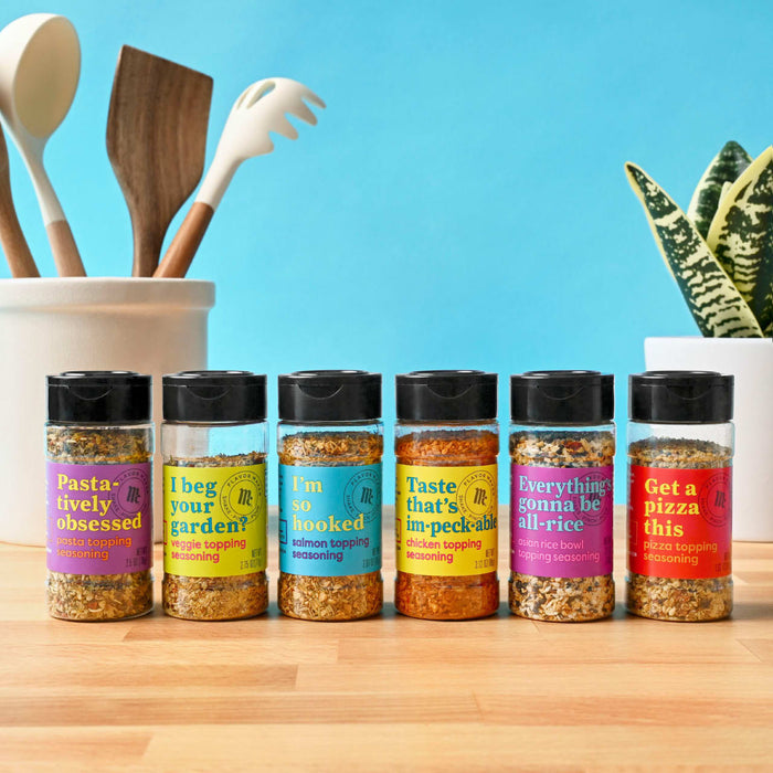 Six jars of Flavor Maker topping seasonings are lined up on a wooden surface with kitchen utensils and a plant in the background.