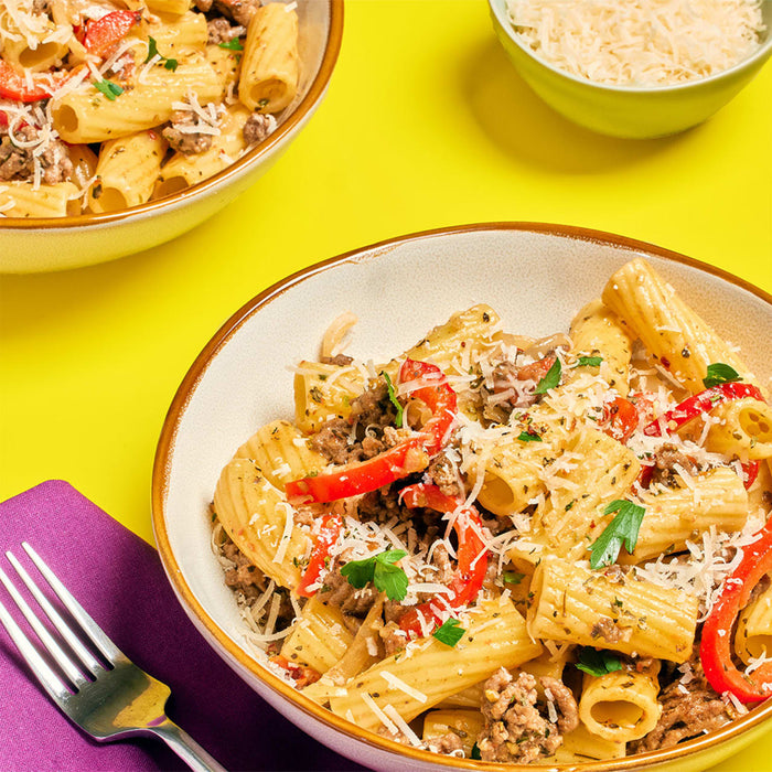 A bowl of rigatoni pasta with meat sauce red peppers and grated cheese is displayed on a yellow background with a fork and a bowl of cheese.