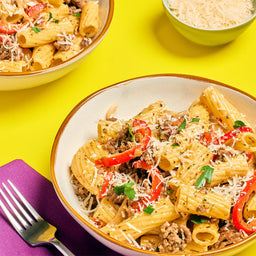 A bowl of rigatoni pasta with meat sauce red peppers and grated cheese is displayed on a yellow background with a fork and a bowl of cheese.