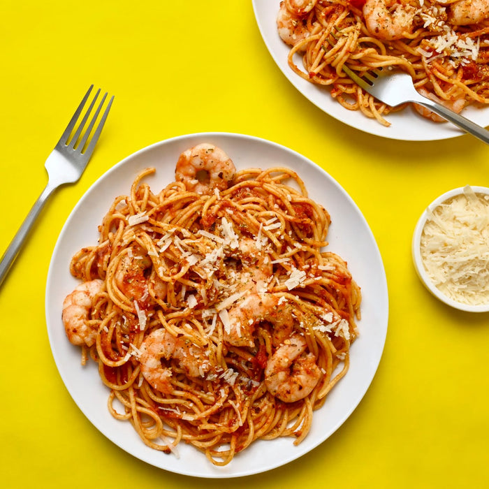 Two plates of shrimp spaghetti with tomato sauce and parmesan cheese are displayed on a vibrant yellow background accompanied by a fork and a bowl of grated cheese.