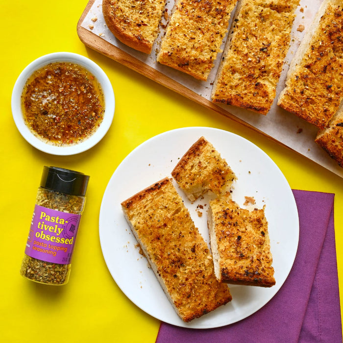 This image features garlic bread seasoned with Flavor Me Pasta-tively Obsessed pasta topping seasoning along with a dipping sauce on a vibrant yellow background.