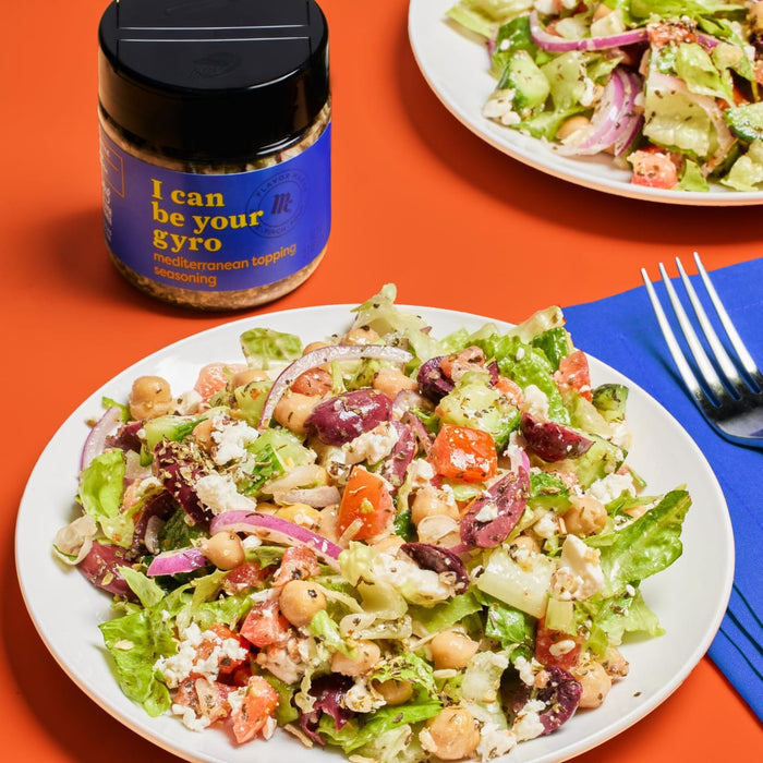 A bowl of salad with chickpeas, feta cheese, tomatoes, red onion, and olives, next to a jar of Mrs. Pinch Flavor Maker I Can Be Your Gyro Mediterranean Topping Seasoning.