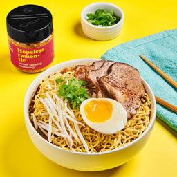 A bowl of ramen noodles with pork, a soft boiled egg, bean sprouts, and green onions, with a jar of Hopeless Ramen-tic seasoning in the background.