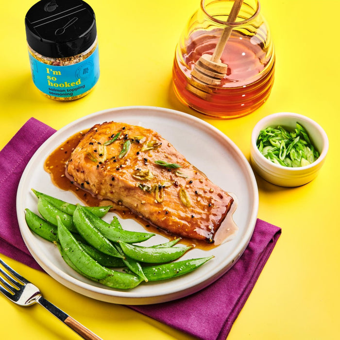 A plate of salmon with green beans and a small bowl of green onions, with a jar of I'm So Hooked salmon topping seasoning and a jar of honey in the background.
