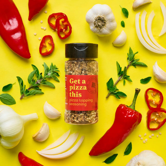 A jar of McCormick Get a Pizza This pizza topping seasoning is surrounded by fresh ingredients like garlic peppers and herbs on a yellow background.