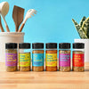 This image shows six Flavor Maker seasoning bottles with different flavors including pasta veggie salmon chicken asian rice bowl and pizza arranged on a wooden surface against a blue background with kitchen utensils and a plant.