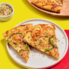 Two slices of pizza with tomatoes green peppers and red onions are displayed on a white plate accompanied by a bowl of seasoning and another pizza slice on a wooden board.