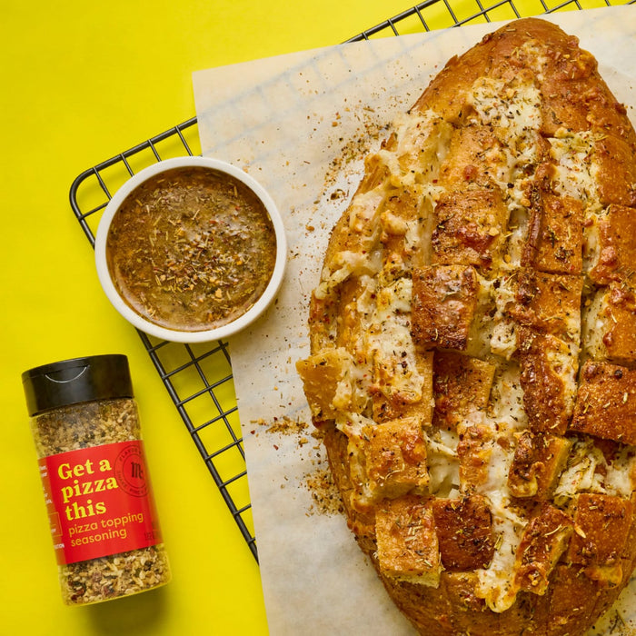 A loaf of cheesy pull-apart bread with a bowl of dipping sauce and a bottle of Flavor Maker Get a pizza this pizza topping seasoning are displayed on a yellow background.