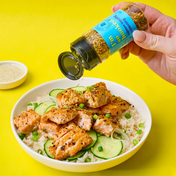 A hand sprinkles Im so hooked salmon topping seasoning onto a bowl of salmon rice and cucumber.