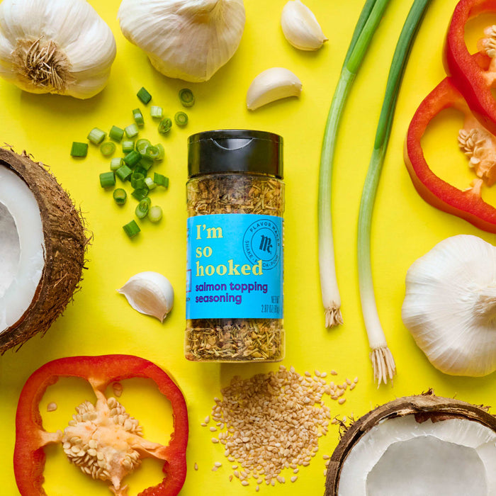 A jar of McCormick Flavor Maker Im So Hooked Salmon Topping Seasoning is surrounded by coconuts garlic green onions red bell pepper and sesame seeds on a yellow background.
