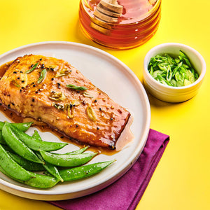 A plate of glazed salmon with snap peas a bowl of chopped scallions and a jar of honey are arranged on a yellow surface.