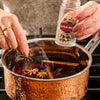 A person adds star anise from a jar of Mccormick Gourmet, 0.42 Oz into a hammered copper pot of simmering mulled wine with cinnamon sticks.