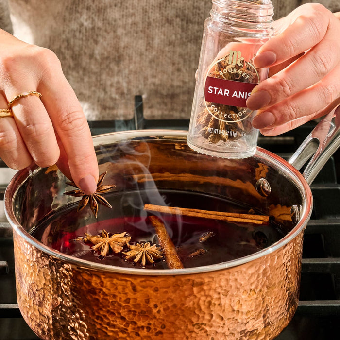 A person adds star anise from a jar of Mccormick Gourmet, 0.42 Oz into a hammered copper pot of simmering mulled wine with cinnamon sticks.