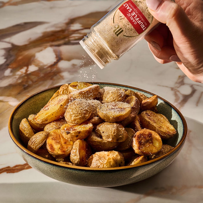 A hand seasons a bowl of golden roasted potatoes by sprinkling them with Mccormick Gourmet Garlic Truffle Salt, 2.25 Oz.