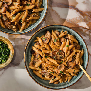 Two bowls of creamy mushroom penne pasta, seasoned with Mccormick Gourmet Garlic Truffle Salt, 2.25 Oz, are served on a marble tabletop with a side of fresh parsley.