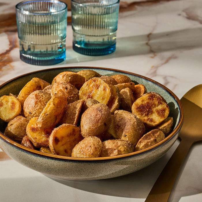 A rustic ceramic bowl filled with golden-brown roasted potatoes, perfectly seasoned with Mccormick Gourmet Garlic Truffle Salt, 2.25 Oz, is presented on a marble surface with a golden spoon and two blue glasses of water in the background.