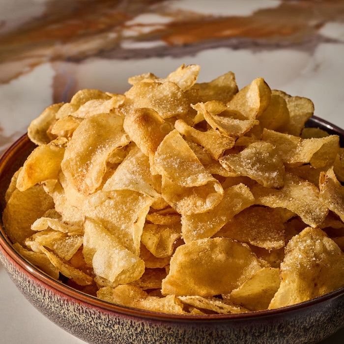 A close-up shot shows a rustic ceramic bowl overflowing with golden potato chips, which have been seasoned with Mccormick Gourmet Garlic Truffle Salt, 2.25 Oz.