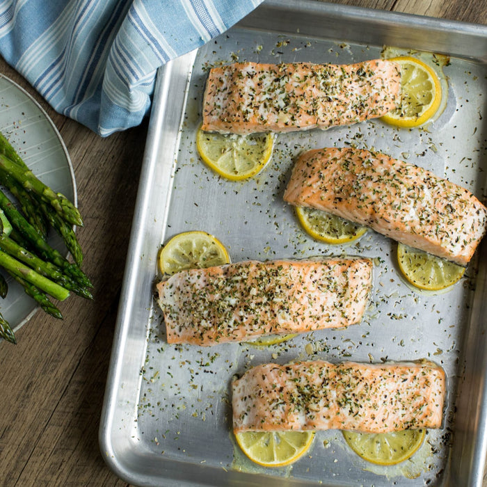 Four salmon fillets, seasoned with herbs and spices, are baked on lemon slices on a metal sheet pan, suggesting preparation with Mccormick Culinary Lemon &amp; Pepper Seasoning Salt, with a side of asparagus and a striped towel.