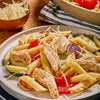 A close-up view of penne pasta with grilled chicken, roasted red bell peppers, zucchini, and red onions, topped with shredded cheese, suggesting a dish seasoned with Mccormick Culinary Lemon &amp; Pepper Seasoning Salt.