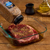 A steak is being seasoned with McCormick Montreal Steak Seasoning in a glass dish.