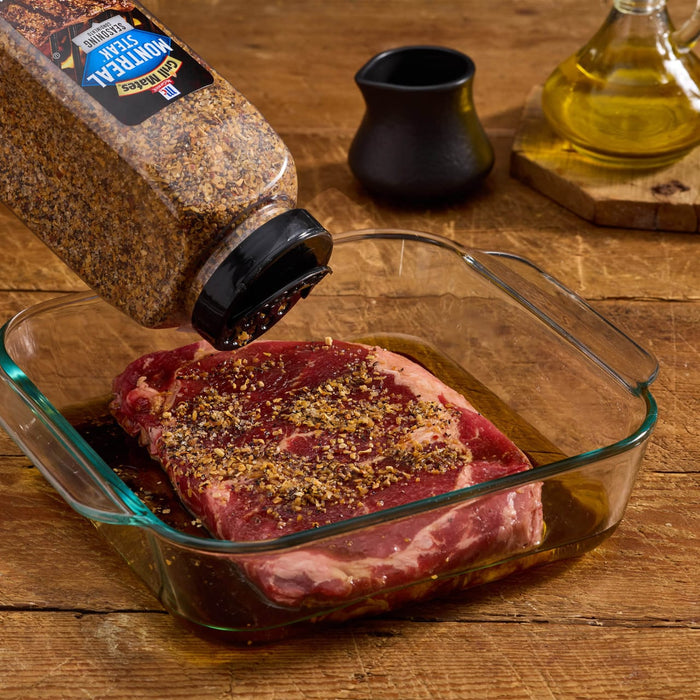 A steak is being seasoned with McCormick Montreal Steak Seasoning in a glass dish.