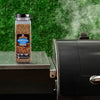 A container of Montreal Steak Spice sits next to a smoking grill, suggesting its use in outdoor cooking.