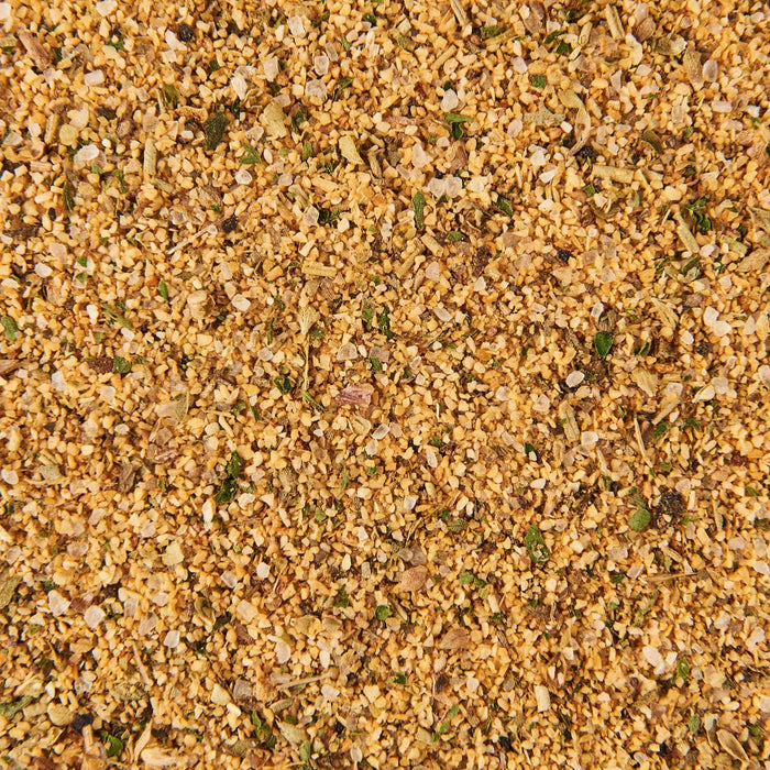A close-up, top-down view of Mccormick Grill Mates Montreal Chicken Seasoning Ground 23 Oz, showing a granular blend of light brown spices, green herbs, and white salt crystals.
