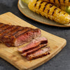 A grilled steak sliced on a wooden board, served with grilled corn on the cob.