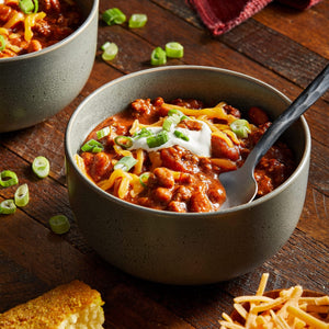 Two bowls of chili topped with shredded cheese, sour cream, and green onions are presented on a wooden surface, suggesting a hearty and comforting meal.