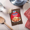 A packet of McCormick Chicken Gravy mix sits on a table next to a wooden spoon and a pot, suggesting a home-cooked meal.